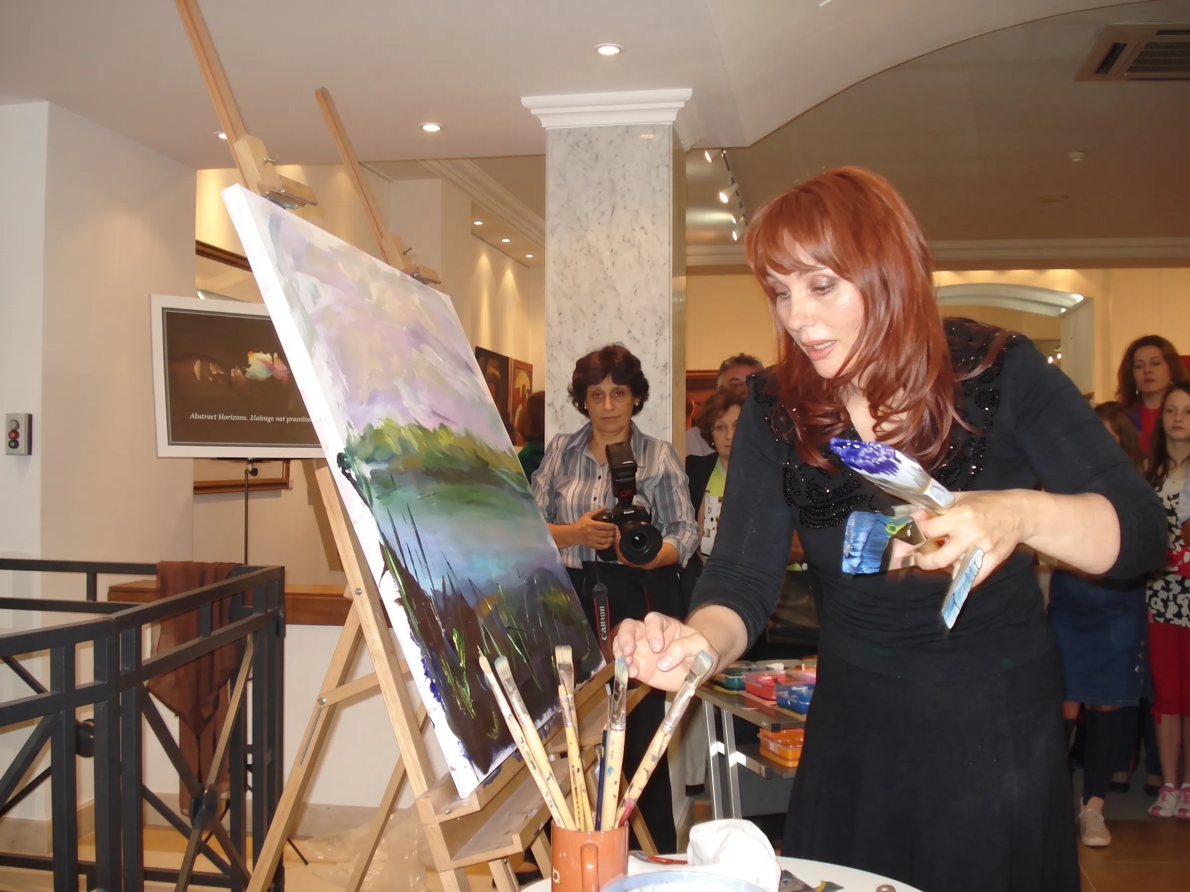 Dinara Dindarova painting at an easel in a gallery, watched by visitors, one of them holding a camera.